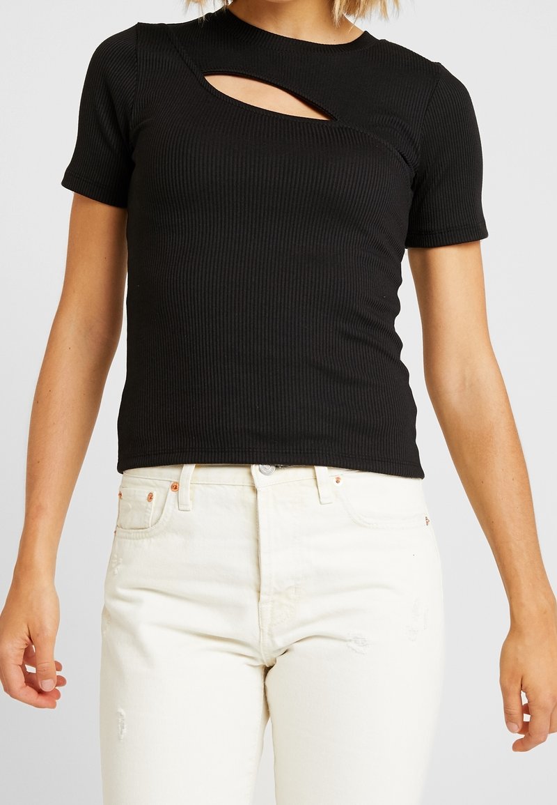 Kvinna iklädd en svart ribbad topp med kort ärm och en utskärning vid halsringningen, tillsammans med högmidjade vita jeans.