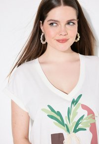 Hvid bomulds-T-shirt med et stort plantegrafik i grønt og brunt. V-halsdesign med rullede ærmer og blød tekstur.
