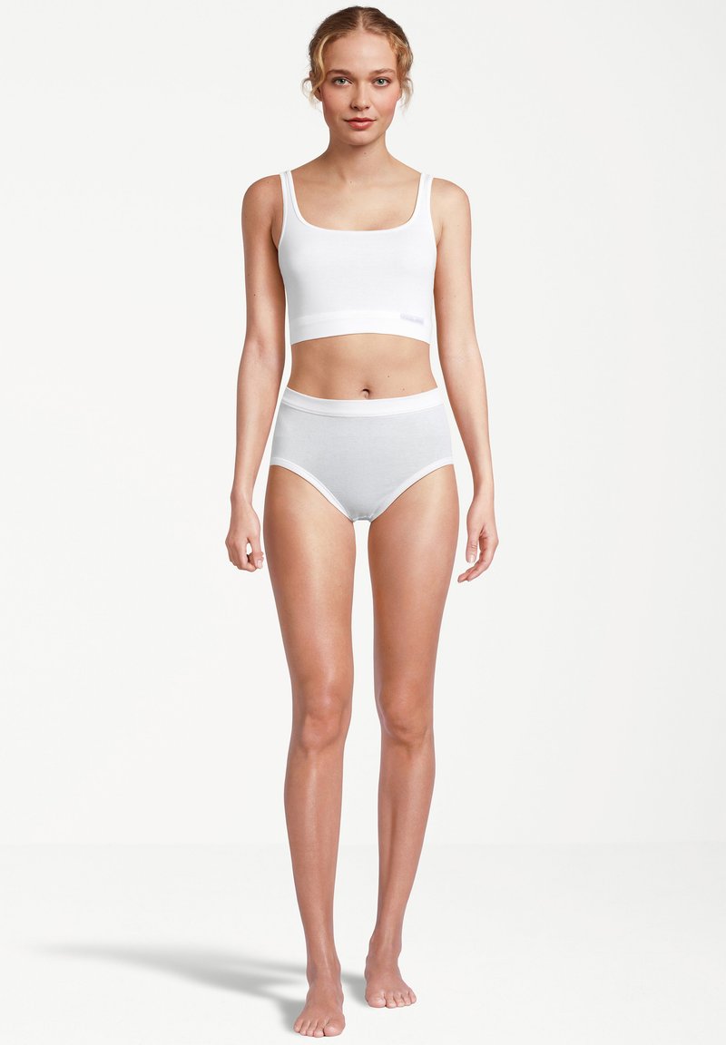 Eine Frau steht barfuß da, trägt ein weißes, kurz geschnittenes Tanktop und hochgeschnittene weiße Unterwäsche vor einem schlichten weißen Hintergrund.