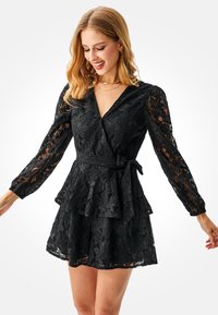 Schwarzes Spitzen-Wickelkleid mit langen Ärmeln, Taillen-Peplum und Bindegürtel. Das Design umfasst florale Muster und einen tiefen V-Ausschnitt.