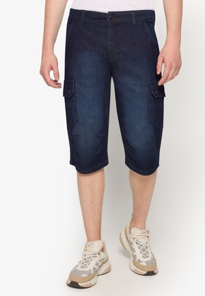 Mørkeblå denim cargoshorts med knaplukning på sidelommer, båret sammen med beige og hvide sneakers på en stående person.