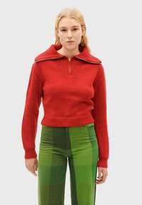 Pull en maille rouge à col large et fermeture éclair sur le devant, avec une texture côtelée et une coupe courte, associé à un pantalon à carreaux verts.