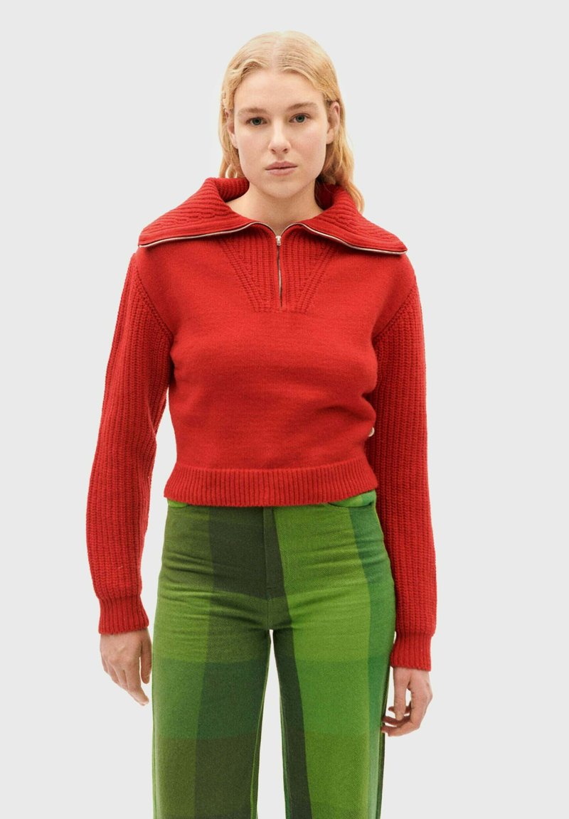Pull en maille rouge à col large et fermeture éclair sur le devant, avec une texture côtelée et une coupe courte, associé à un pantalon à carreaux verts.