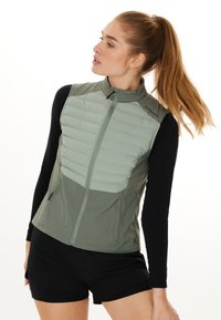 Leichter, grüner Steppwesten mit horizontaler Steppung, hohem Kragen und Reißverschluss, kombiniert mit einem schwarzen Langarmoberteil und Shorts.