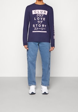 Tummansininen pitkähihaisessa paidassa on vaaleanpunainen graafinen teksti, joka on yhdistetty vaaleansiinisiin farkkuihin. Kengät ovat valkoiset mustilla yksityiskohdilla. Yksinkertainen, rento tyyli.