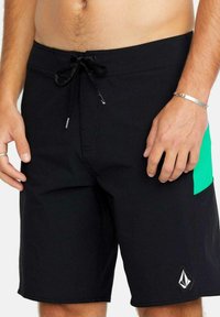 Bañador negro con un panel lateral verde, cintura con cordón y un logo de la marca en blanco. Fabricado en tejido ligero y de secado rápido.