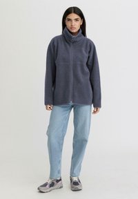 Giacca in pile grigio scuro con zip e colletto alto, caratterizzata da una texture morbida e due tasche laterali, abbinata a jeans in denim chiaro e scarpe da ginnastica.