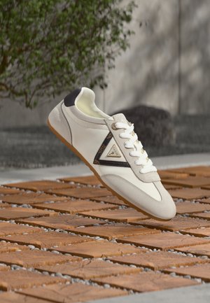 Baskets blanches avec détails en daim, patch noir au talon et logo triangulaire sur le côté, présentées sur un pavé bois et pierre texturé.