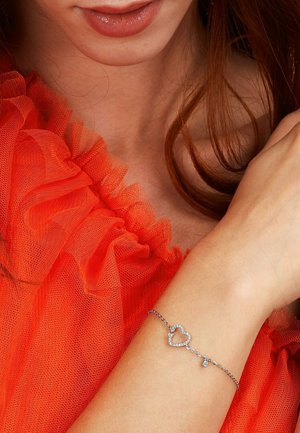 Woman wearing a silver heart-shaped charm bracelet on her wrist and a bright orange ruffled top, with loose auburn hair.