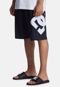 Schwarze Shorts mit einem großen weißen Grafikdesign an der Seite, kombiniert mit schwarzen Rutschen-Sandalen, die ein weißes Logo tragen. Das Material scheint leicht und flexibel zu sein.