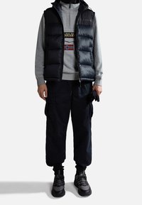 Gilet imbottito nero sopra una felpa grigia con un design stampato, abbinato a pantaloni cargo neri e sneakers grigio-neri.