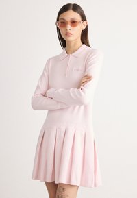 Ružová polokošeľa s dlhým rukávom a plisovanou sukňou, vyrobená z mäkkého materiálu, s golierom a malým logom na hrudi.