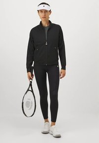 Sort atletikjakke, tætsiddende sorte leggings, hvid kasket og hvide sneakers. Holder en tennisketcher, minimalistisk design, tekstureret materiale.