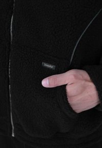 Mano parzialmente all'interno della tasca di una giacca in pile nera con cerniera visibile e piccolo etichetta del marchio sulla tasca.