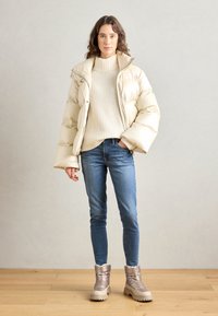 Doudoune beige avec des manches larges, col roulé crème côtelé, jeans skinny bleus et bottes d'hiver métalliques taupe avec fausse fourrure.