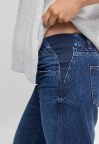 Eine Hand hebt einen grauen Pullover an, um ein marineblaues elastisches Panel an der Taille einer blauen Umstandsjeans zu zeigen.