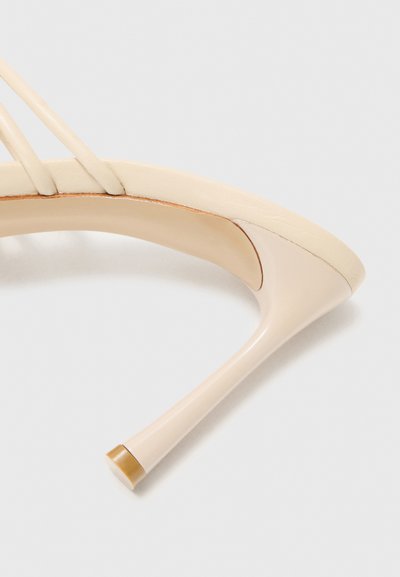 Sandalia de tacón alto beige con tiras finas y un tacón de aguja delgado sobre un fondo blanco.