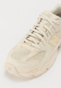 Baskets beige New Balance avec des empiècements en mesh et synthétiques, lacets beige et logo de la marque sur le côté et sur la languette.