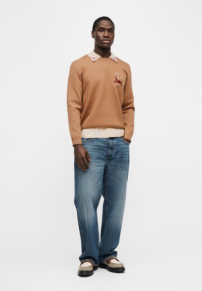 Άνδρας με μπεζ πουλόβερ με κέντημα αλόγου, πουκάμισο με κρεμ γιακά, άνετο μπλε τζιν και διχρωμίες loafers, στέκεται μπροστά σε λευκό φόντο.