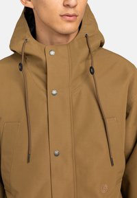 Mann, der eine braune Kapuzenjacke mit Kordeln, metallenen Druckknöpfen und einem kleinen gestickten Logo im Bereich der Vordertasche trägt.