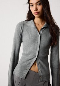 Sivá rebrovaná cardigan so dlhými rukávmi, gombíkmi na prednej strane a špicatým golierom s dekoratívnymi kovovými detailmi. Mäkkučký textura.