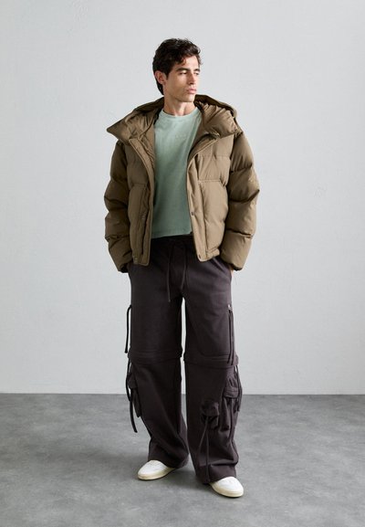 Braune Pufferjacke mit Kapuze und Druckknöpfen über einem hellgrünen Hemd. Dunkle Cargo-Hosen mit mehreren Taschen und Kordelzügen. Weiße Sneakers.