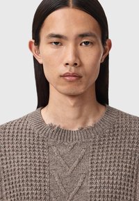 Strikkegenser i lys brun med et teksturert mønster, med rund hals, slitte kanter og en kabelstrikkedesign på den ene siden.