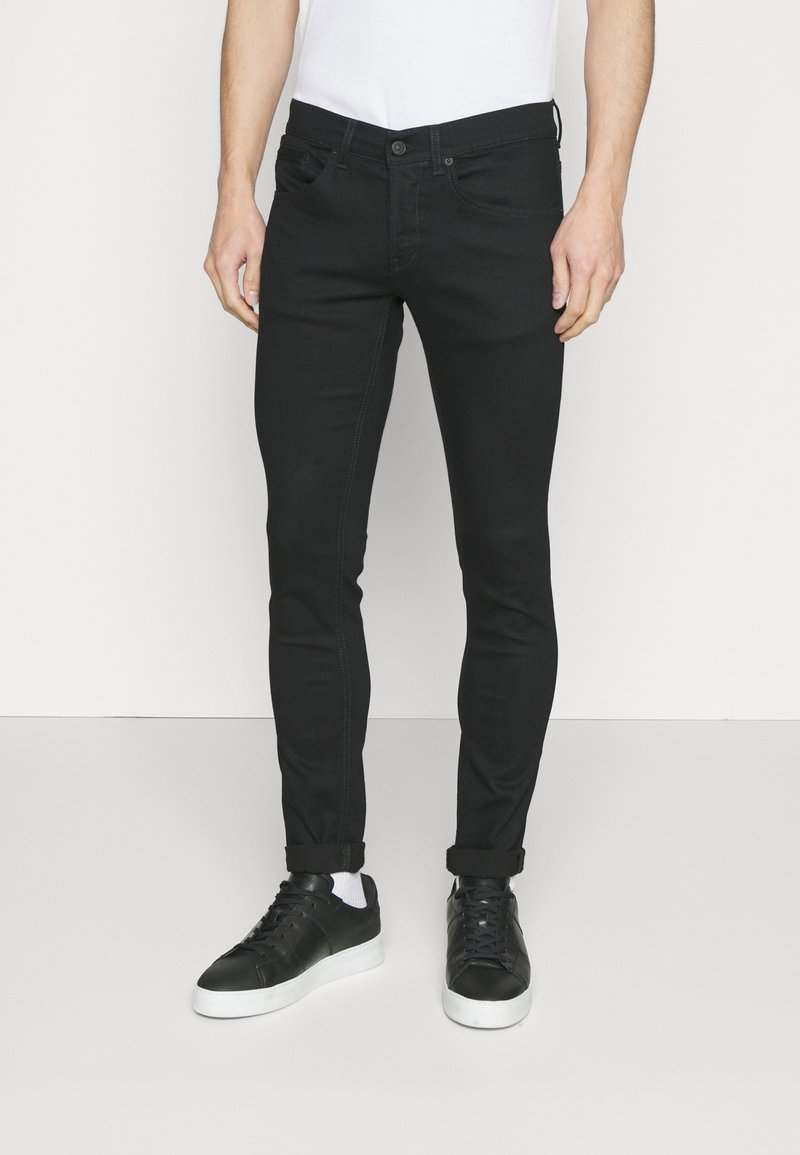 Homme portant un jean slim noir retroussé aux chevilles, associé à des baskets noires à semelles blanches et des chaussettes blanches.