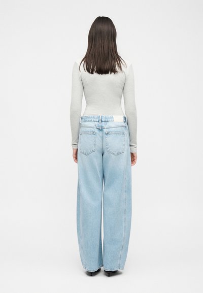 Γυναίκα που στέκεται με την πλάτη, φοράει ανοιχτό γκρι μακρυμάνικη μπλούζα και ανοιχτό μπλε φαρδιά παντελόνια τζιν σε λευκό φόντο.