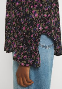 Svart blommig blus med små rosa och blå blommor, samlade ärmar och elastiska ärmslut, kombinerad med ljusblå denimjeans.