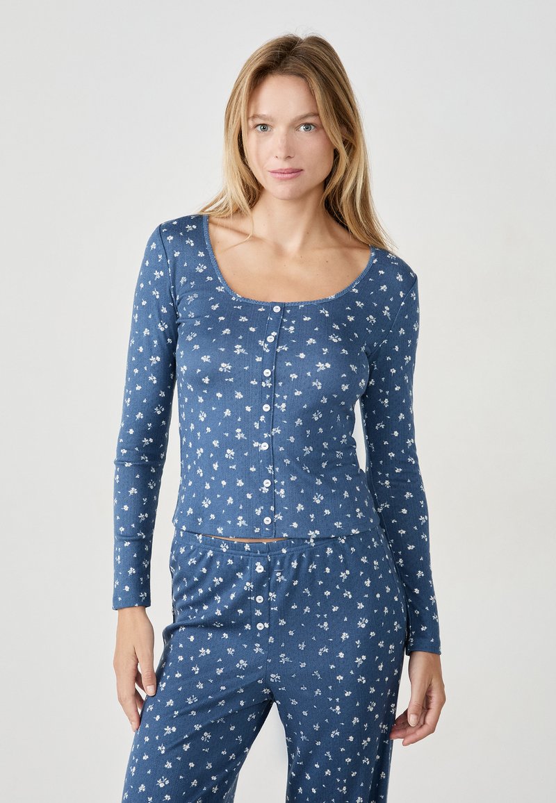 Kobieta w niebieskiej, długiej bluzce z guzikami i dopasowanych spodniach w drobny biały kwiatowy wzór, stojąca na jednolitym jasnym tle.