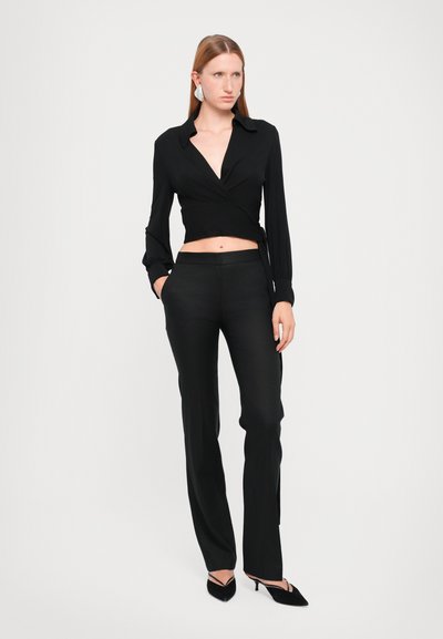 Blouse noire à manches longues, associée à un pantalon noir cintré. Les deux sont en tissu lisse, présentant un design ajusté et une fente sur le côté.