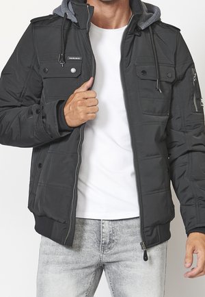 Mann trägt schwarze Kapuzenjacke über weißem Hemd und hellgrauen Jeans, teilweise den Reißverschluss der Jacke schließend.