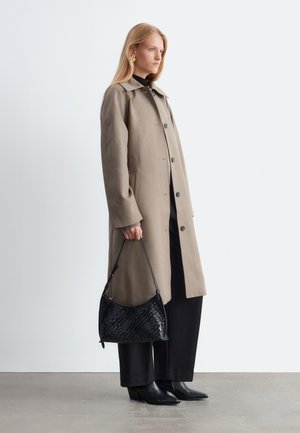 Femme portant un long manteau beige, un pantalon noir, des bottes noires et des boucles d'oreilles dorées, tenant un sac à main noir texturé dans sa main gauche.