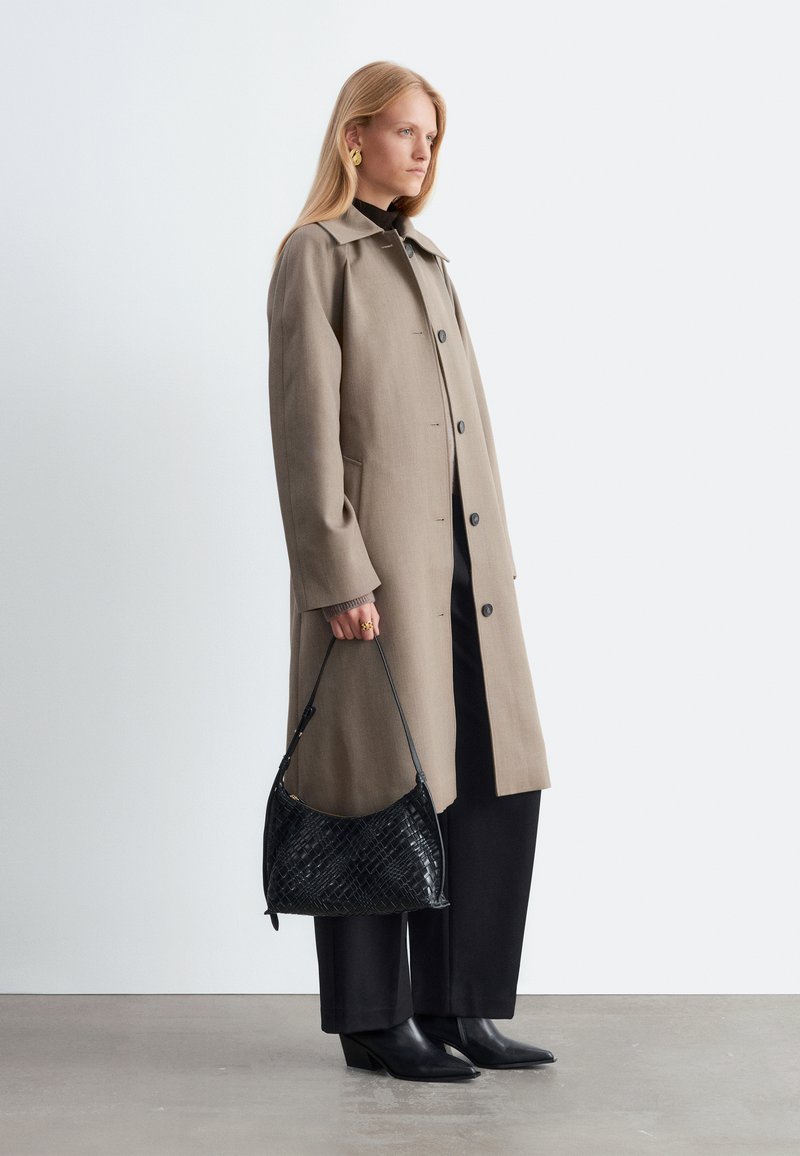 Femme portant un long manteau beige, un pantalon noir, des bottes noires et des boucles d'oreilles dorées, tenant un sac à main noir texturé dans sa main gauche.
