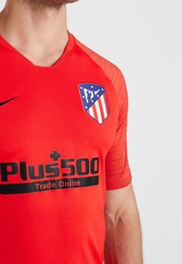 Camiseta de fútbol roja del Atlético de Madrid con el escudo del club en el pecho y el logo del patrocinador Plus500 Trade Online en negro.