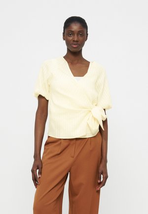 Žena u blijedo žutoj bluzi na preklop s mašnom i smeđim širokim hlačama, stoji na jednostavnoj svijetloj pozadini.