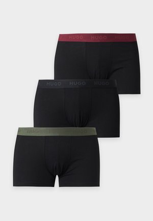 Ung mand, der står barfodet iført sorte boxershorts med en mørkerød linning mod en ensfarvet lys baggrund.