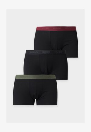 Jeune homme debout, pieds nus, portant un boxer noir avec une ceinture rouge foncé sur un fond clair uni.