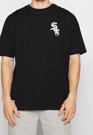 Mężczyzna w czarnej koszulce z krótkim rękawem z białym logo "Sox" na lewej piersi oraz w jasnoszarych spodniach dresowych na jednolitym tle.