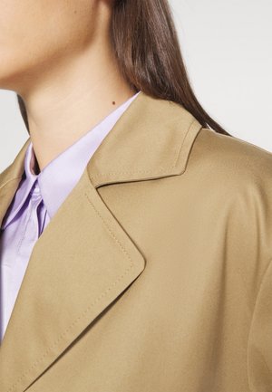 Mujer con abrigo tipo trench beige sobre camisa lavanda con cuello, mostrando detalles del escote y los hombros.