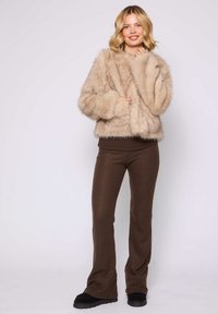 Beige Kunstfelljacke mit runder Form, weiten Ärmeln und geripptem Saum. Kombiniert mit dunkelbraunen Schlaghosen und schwarzem Schuhwerk.