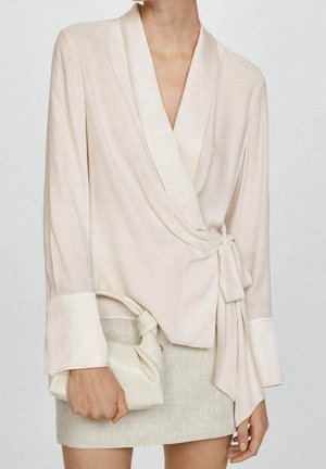 Mujer que lleva una blusa cruzada de color crema con puños anchos y sostiene un pequeño bolso a juego en color crema, combinada con una falda beige claro.