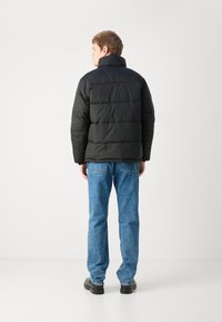 Zwarte pufferjack met horizontaal gestikte secties, hoge kraag en een relaxte pasvorm, gedragen met lichtblauwe spijkerbroeken en zwarte schoenen.