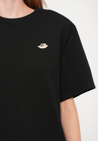 Black cotton t-shirt with short sleeves and a round neck. Features a small embroidered design on the chest depicting a pair of wings.