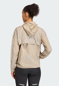 Beige lett jakke med hette, med et semi-transparent mønster på yoken, elastiske ermer og reflekterende detaljer på baksiden.