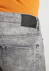 Lähikuva harmaista happopesu farkuista, takataskusta ja mustasta bränditunnuksesta, joita on yhdistetty oranssiin toppiin.