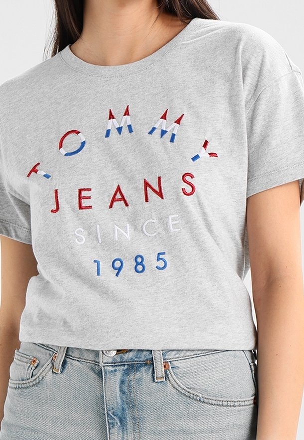 Kobieta ubrana w jasnoszary T-shirt z napisem "TOMMY JEANS SINCE 1985" w czerwonych, białych i niebieskich literach, zestawiony z jasnoniebieskimi dżinsami.