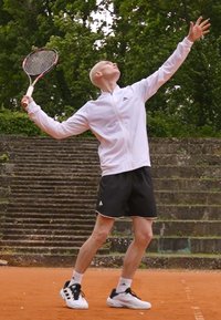 Jucător de tenis îmbrăcat într-o jachetă albă cu fermoar și pantaloni scurți negri, ținând o rachetă, pe fundalul unui peisaj natural cu trepte de piatră și vegetație.