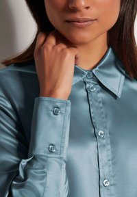 Hellblaues Satinhemd mit spitzem Kragen, knöpfbarem Vorderteil und langen Ärmeln mit Knopfbündchen. Glatte Textur und subtiler Glanz.